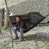 A person sitting in a DD Nest Hammock with a DD Multicam pattern, camouflaged under a canopy in a woodland setting.